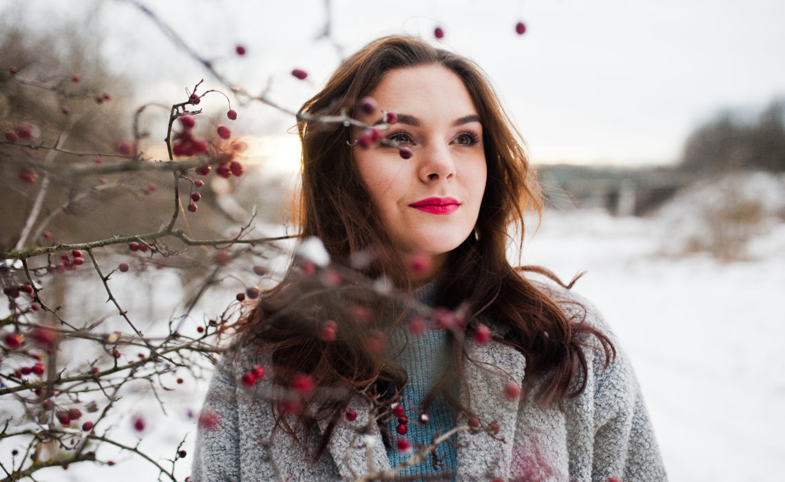 Close up portrait of gentle girl in gray coat near the branches of a snow-covered tree.