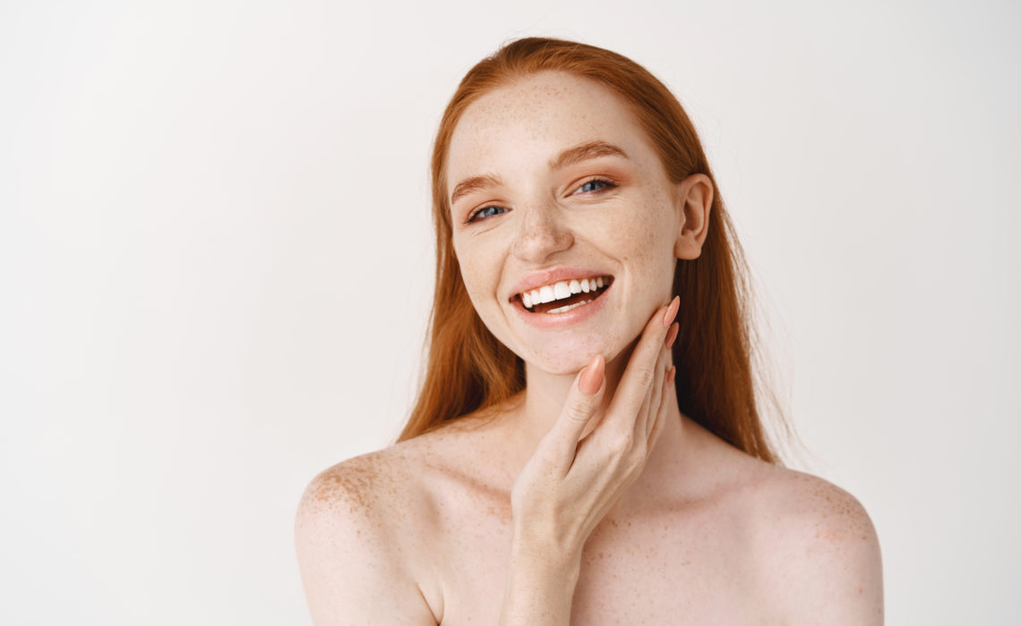 Beauty. Close-up of young beautiful redhead woman smiling at camera, touching perfect clean skin on face and looking happy, standing naked over white background.