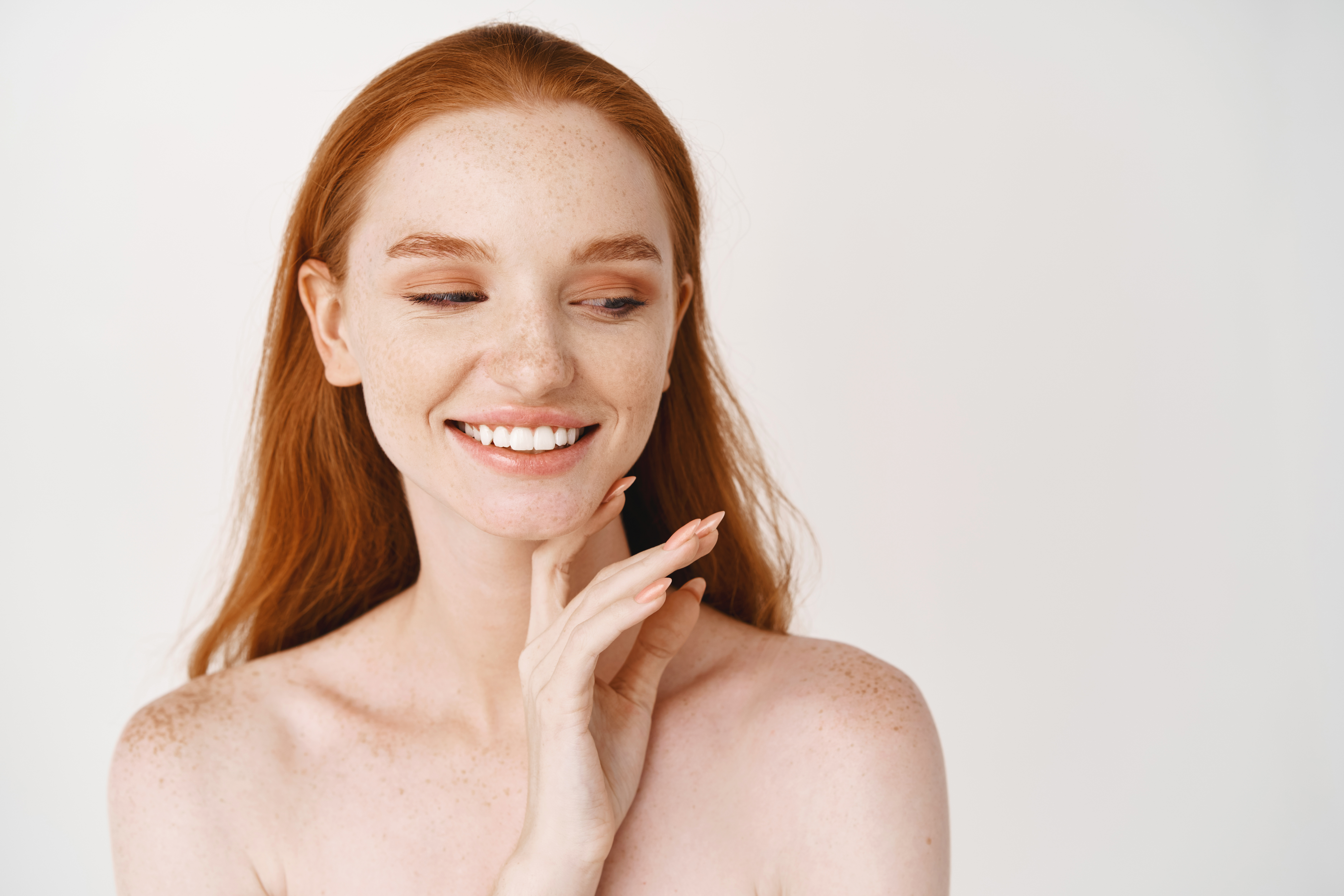 Skincare. Close-up of redhead woman with pale soft skin, smiling white teeth and touching clean no makeup face, looking aside, standing over white background naked.